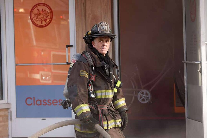 Christopher Herrmann wears full firefighting gear and holds a hose near the entrance of a building in Chicago Fire Episode 1202.