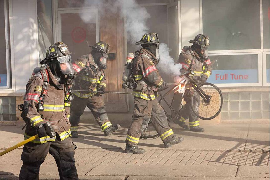 Smokey firefighters walk on a sidewalk holding tools and a scorched bike in Chicago Fire Episode 1202.