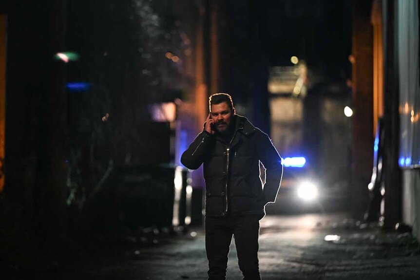 Adam Ruzek speaks on a cellphone in an alley as a police car drives in the background in Chicago P.D. Episode 1102