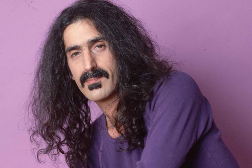 Frank Zappa wears a purple shirt in front of a purple background.